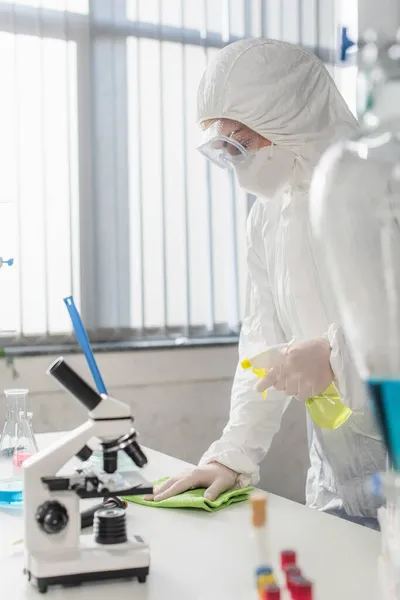 Doctor in hazmat suit cleaning table near microscope in lab — Stock Photo