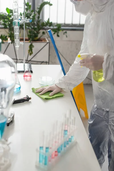 Wissenschaftler im Schutzanzug wischt Schreibtisch mit Lappen ab, während er Sprühflasche hält — Stockfoto