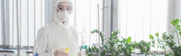 Scientist in hazmat suit looking at camera while holding spray bottle, banner — Stock Photo