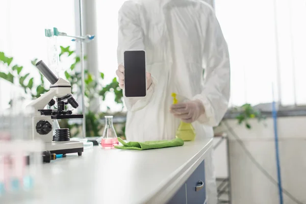 Vue recadrée du médecin montrant smartphone avec écran blanc près du flacon et microscope sur le bureau — Photo de stock