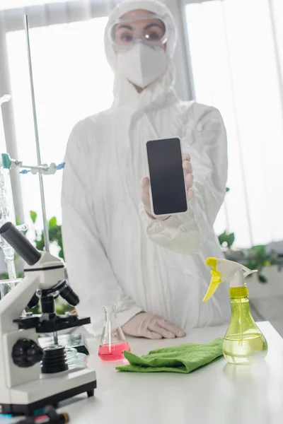 Scientifique flou dans l'équipement de protection individuelle montrant smartphone avec écran blanc près du microscope en laboratoire — Photo de stock