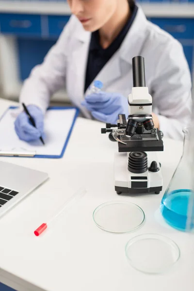 Vista ritagliata della donna sfocata che scrive sugli appunti vicino al microscopio e sulle stoviglie di Petri sulla scrivania — Foto stock