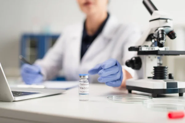 Partial view of blurred doctor near microscope and coronavirus vaccine — Stock Photo