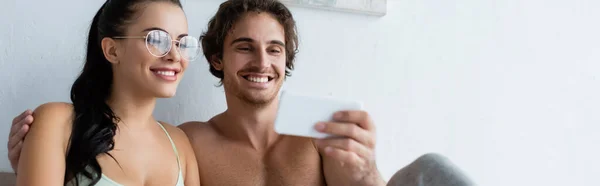 Shirtless man holding blurred smartphone near smiling girlfriend in eyeglasses at home, banner — Stock Photo