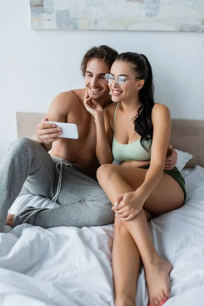 Femme souriante dans des lunettes touchant sans chemise copain prendre selfie sur smartphone sur le lit — Photo de stock