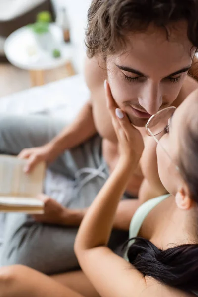 Vista de ángulo alto de la mujer en anteojos besándose hombre sin camisa con libro borroso en la cama - foto de stock