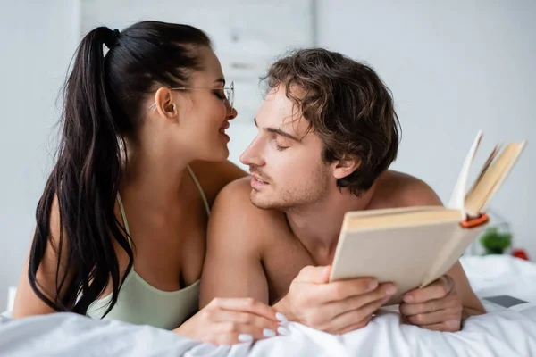 Femme gaie dans les lunettes couché près du petit ami avec livre sur le lit — Photo de stock