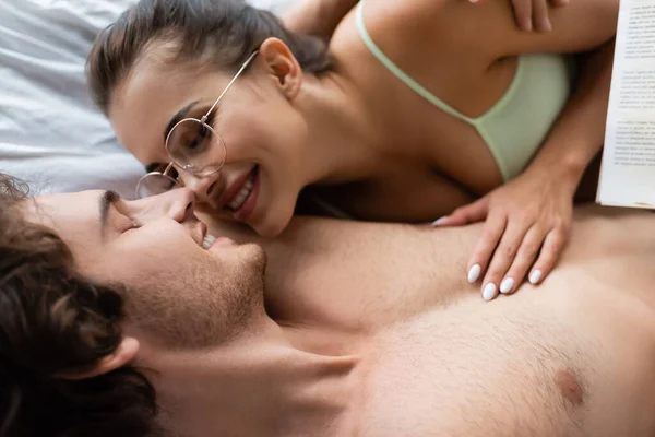 Visão superior da jovem mulher em óculos sorrindo ao tocar namorado perto do livro na cama — Fotografia de Stock