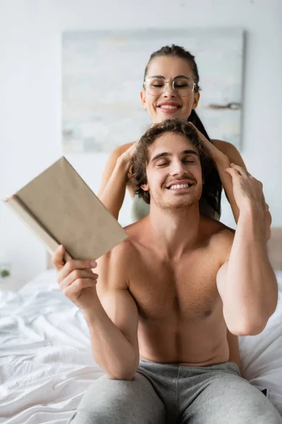 Улыбающаяся женщина в очках трогает голову бойфренда без рубашки держа книгу на кровати — стоковое фото