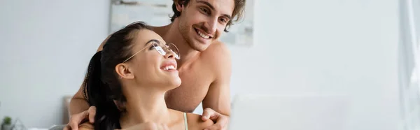 Shirtless man hugging girlfriend near blurred laptop at home, banner — Stock Photo