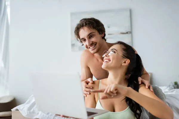 Femme souriante dans les lunettes pointant vers un ordinateur portable près de copain torse nu sur le lit — Photo de stock