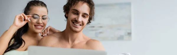 Hombre sin camisa mirando a la computadora portátil cerca de la novia sonriente en gafas en casa, pancarta - foto de stock