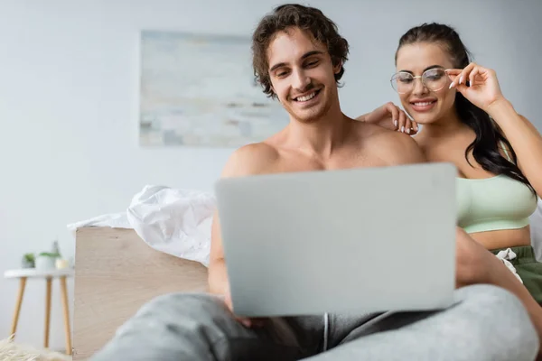 Femme sexy souriante dans les lunettes de vue en regardant ordinateur portable flou près de copain torse nu dans la chambre — Photo de stock