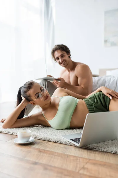 Mujer sexy mirando portátil cerca de café y novio borroso en el dormitorio - foto de stock