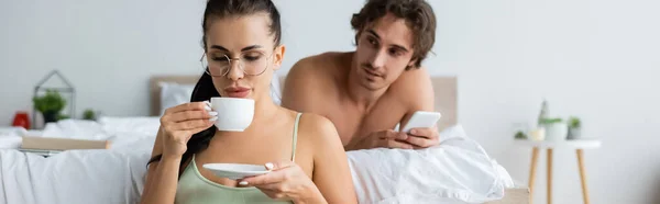 Uomo senza maglietta utilizzando smartphone vicino ragazza sexy bere caffè in camera da letto, banner — Foto stock