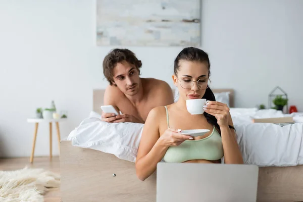 Freelancer sexy segurando café perto de laptop e namorado sem camisa com smartphone na cama — Fotografia de Stock