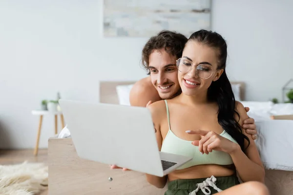 Donna sorridente in alto e occhiali che puntano al computer portatile vicino al fidanzato senza maglietta in camera da letto — Foto stock