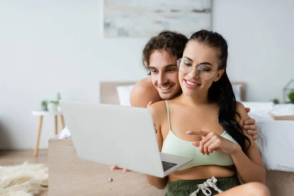 Smiling woman in top and eyeglasses pointing at laptop near shirtless boyfriend in bedroom — Stock Photo