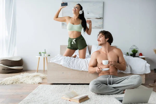 Shirtless man holding cup near laptop, book and smiling girlfriend in headphones in bedroom — Stock Photo
