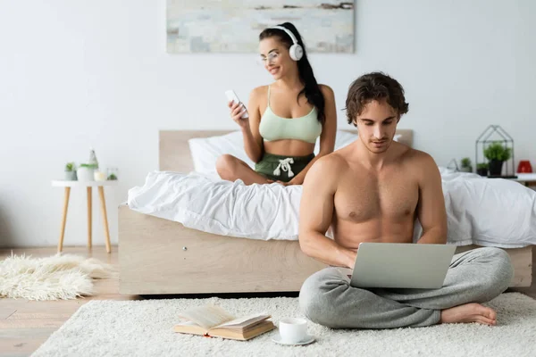 Мужчина без рубашки, пользующийся ноутбуком рядом с кофе и размытой девушкой со смартфоном и наушниками на кровати — стоковое фото