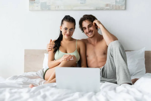 Smiling man hugging sexy girlfriend near blurred laptop on bed — Stock Photo