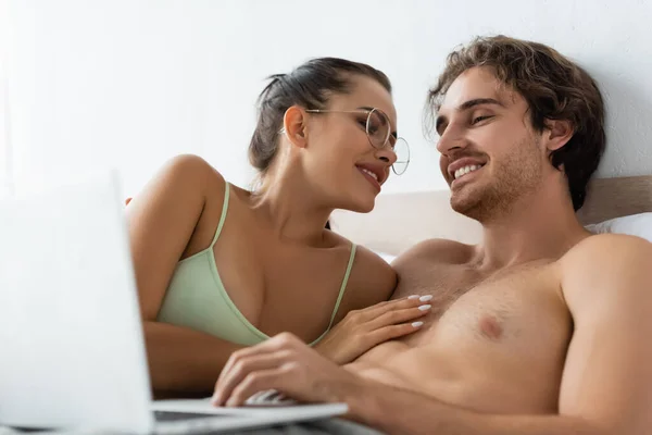 Mulher sexy tocando namorado sem camisa usando laptop desfocado na cama — Fotografia de Stock