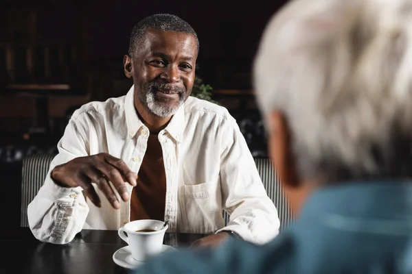 Улыбающийся старший африканский американец смотрит на размытого друга возле чашки кофе — стоковое фото