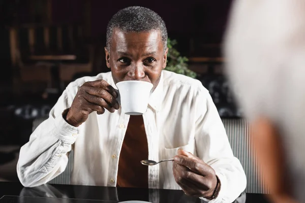 Anziano africano americano uomo bere caffè vicino offuscata amico in caffè — Foto stock