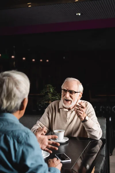 Senior mit Brille gestikuliert im Gespräch mit verschwommenem Freund im Café — Stockfoto