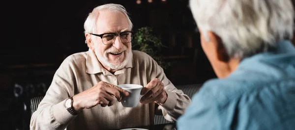 Feliz hombre mayor en anteojos sosteniendo taza de café cerca de amigo borroso en la cafetería, pancarta — Stock Photo