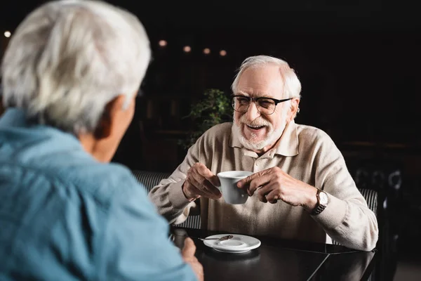 Gioioso uomo anziano in occhiali in possesso di tazza di caffè vicino amico offuscata nel bar — Foto stock