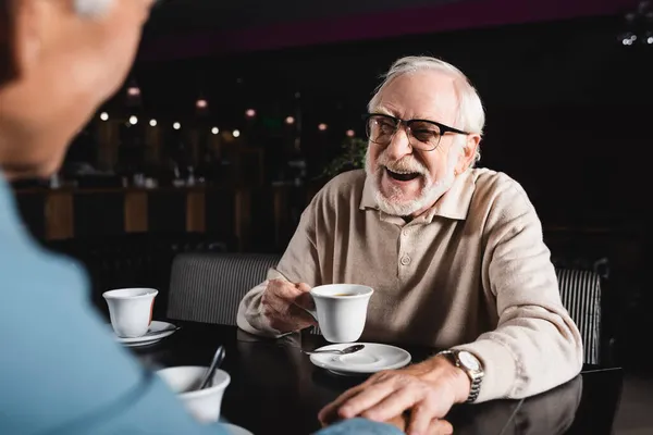 Cheerful senior man in eyeglasses holding cup of coffee while touching hand of blurred friend in cafe — Stock Photo