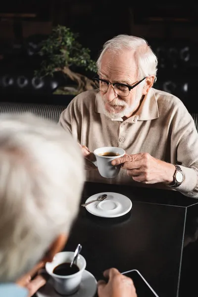 Пожилой человек в очках держит чашку кофе рядом с размытым другом в кафе — стоковое фото
