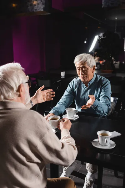 Старші міжрасові друзі жестикулюють під час розмови біля чашок кави — стокове фото