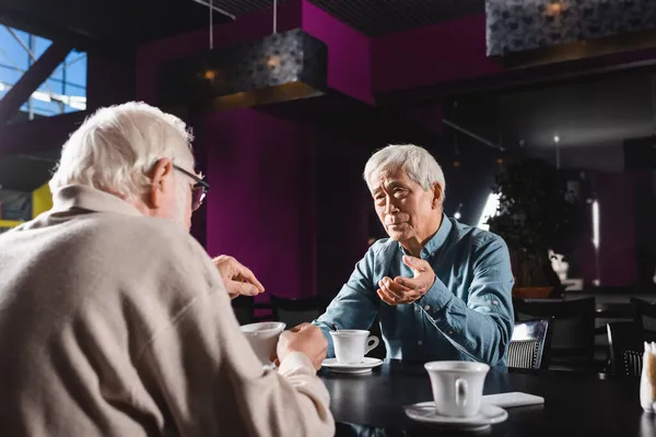 Senior men gesturing while talking with asian friend in cafe near cups of coffee — Stock Photo