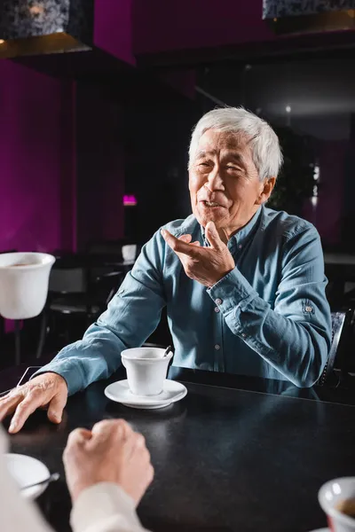 Positivo asiático sênior homem gestos enquanto falando para desfocado amigo no café — Fotografia de Stock