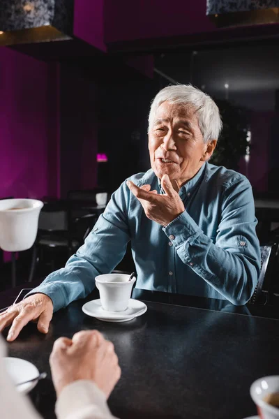 Positive asian senior man gesturing while talking to blurred friend in cafe — Stock Photo