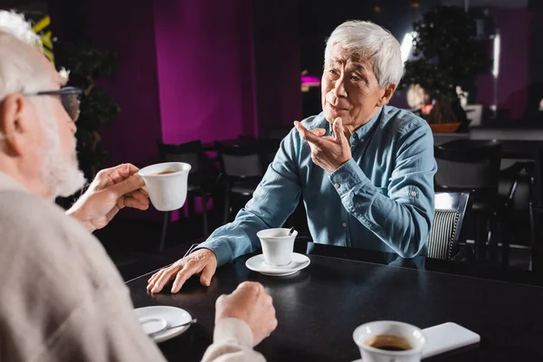 Borrosa hombre celebración taza de café cerca senior asiático hombre gesto mientras hablando en café - foto de stock
