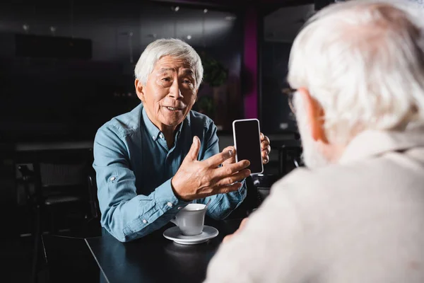 Senior asian man showing smartphone with blank screen to blurred friend in cafe — Stock Photo
