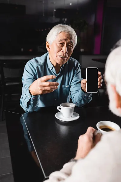 Senior asiático hombre apuntando a celular con blanco pantalla mientras pasar tiempo con borrosa amigo en café - foto de stock