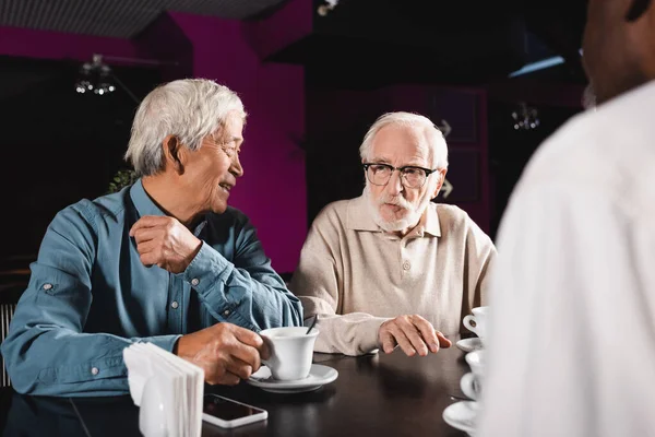 Smiling asian man talking to senior multiethnic friends in cafe — Stock Photo