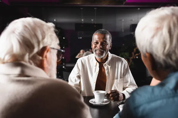 Happy african american man sitting in bar near cup of coffee and blurred senior friends — Stock Photo