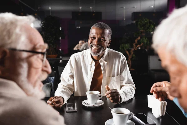 Älterer afrikanisch-amerikanischer Mann lächelt in der Nähe verschwommener multiethnischer Menschen, die im Café reden — Stockfoto