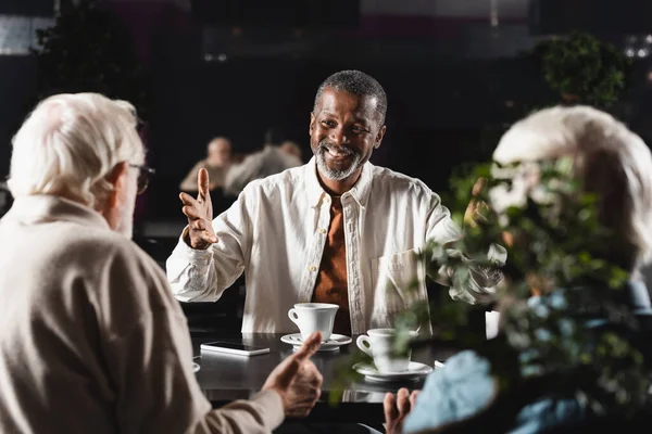 Happy african american man pointing with hands at blurred senior friends in cafe — Stock Photo