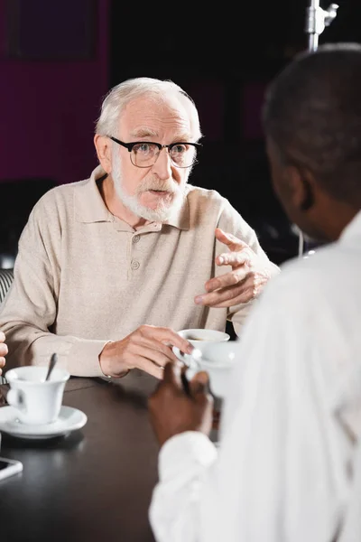Senior man in eyeglasses gesturing while talking to blurred african american man in cafe — Stock Photo