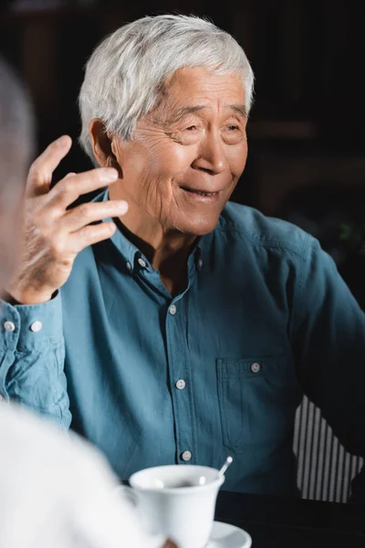 Positive senior asian man gesturing while talking to blurred african american friend in bar — Stock Photo