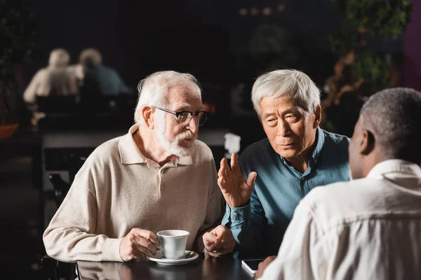 Elderly asian man talking and gesturing near multiethnic friends in cafe — Stock Photo