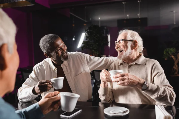 Uomo anziano in occhiali da vista ridere mentre beve caffè con amici multiculturali in caffè — Foto stock