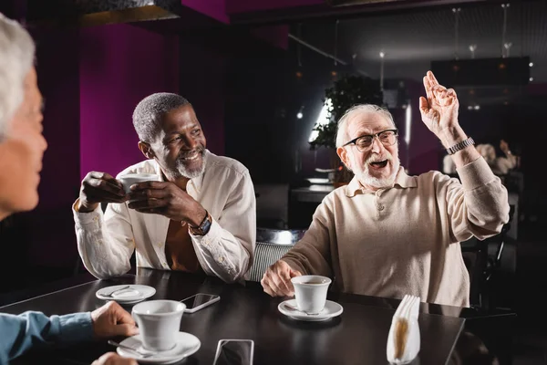 Amazed senior man looking away and waving hand while spending time with multiethnic friends in cafe — Stock Photo