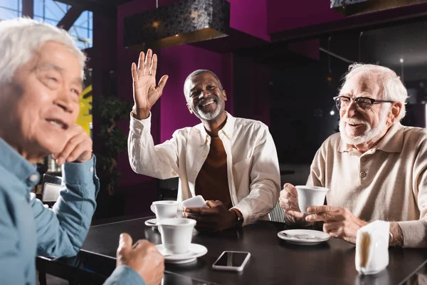 Allegro uomo afro-americano che distoglie lo sguardo e agita la mano vicino agli amici multietnici anziani nel caffè — Foto stock